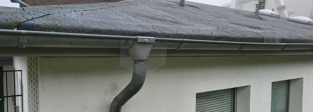 Das Bild zeigt ein weißes Wohnhaus mit Flachdach, an dessen Fallrohr eine transparente Mardersperre montiert ist. Diese Vorrichtung blockiert den Aufstieg von Mardern, Katzen oder anderen Tieren und schützt so Dach und Dachboden vor ungebetenen Gästen. Dank des klaren Materials bleibt die Sperre dezent und fügt sich optisch unauffällig in die Fassade ein. Eine effektive, langlebige und wetterfeste Lösung zur Tierabwehr für Hausbesitzer.